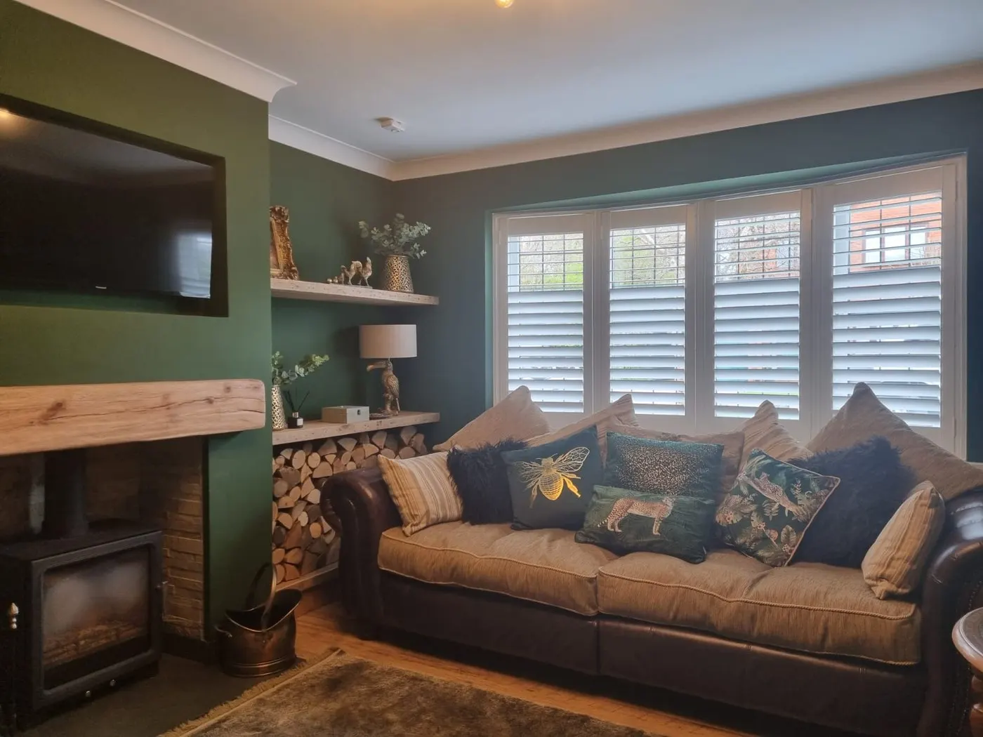 Light grey plantation shutters in a bay window in a green living room with a log burner