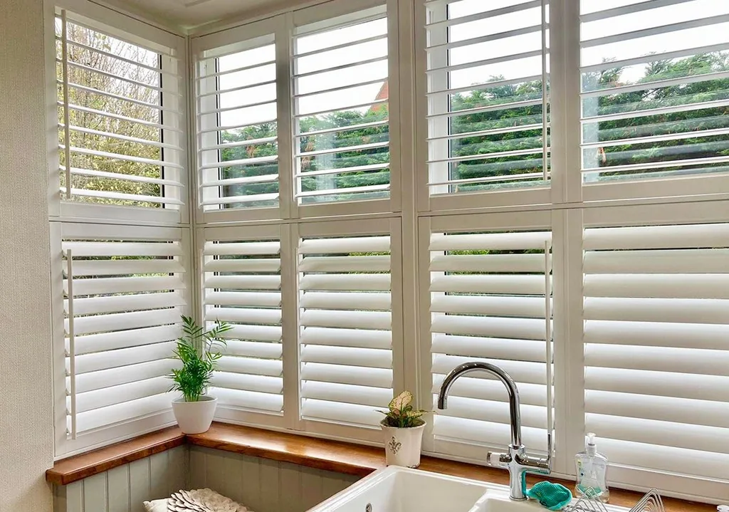 White plantation shutters on a corner kitchen bay window overlooking a leafy garden