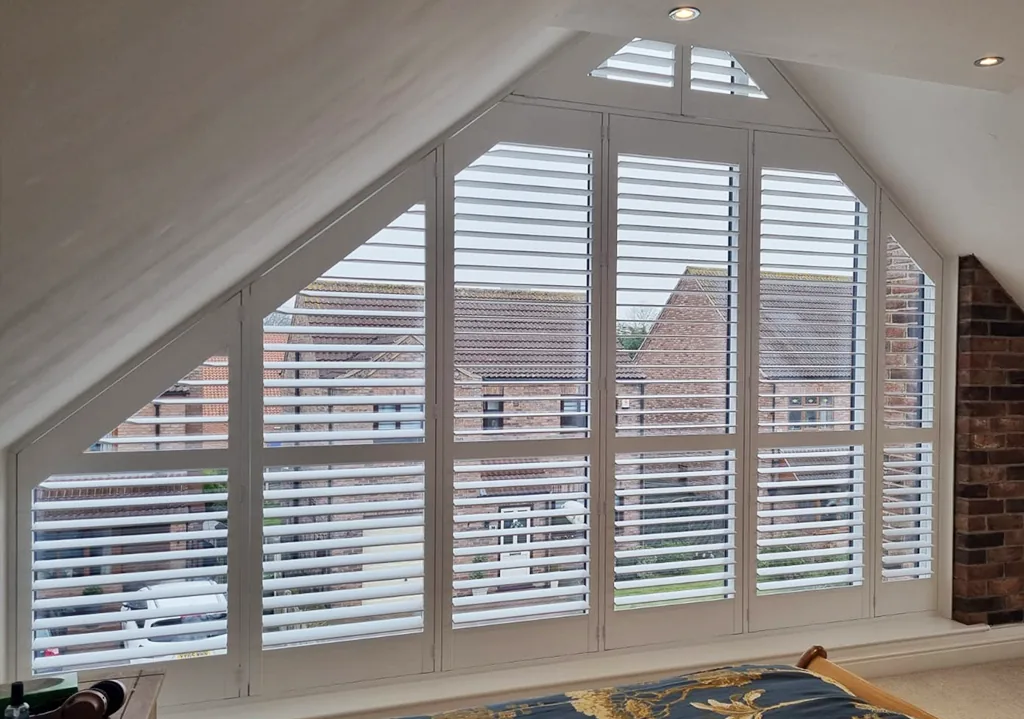 Bespoke white plantation shutters fitted to a shaped apex window in a loft bedroom